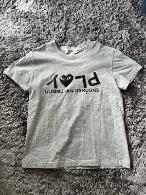 Grå Comme des Garçons PLAY t-shirt - Snygg grå t-shirt från Comme des Garçons PLAY med svart tryck och det ikoniska hjärtögat på bröstet. Klassisk passform med rund hals och korta ärmar. Perfekt till jeans eller shorts för en clean och trendig look. Pris kan diskuteras. 