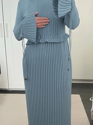 Ljusblå plisserad långklänning - Säljer en elegant ljusblå långklänning med plisserad design och volangdetaljer vid ärmslut och midja. Klänningen har långa ärmar och ett flowy fall som ger en stilren look. Perfekt för dig som gillar en modern och chic stil.Gillar du inte just denna klänning kolla in i min profil och se om du gillar något där!