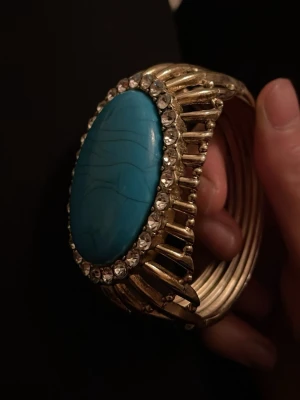 Guldigt armband med blå sten. Det är ett manschettarmband. 😊 - Snyggt armband i guldfärgad metall med en stor oval blå sten i mitten. Runt stenen sitter glittrande klara stenar som ger extra bling. Armbandet har en bred och unik design som verkligen sticker ut. 