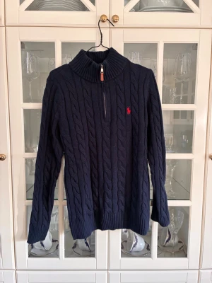 Mörkblå kabelstickad tröja Polo Ralph Lauren - Mörkblå kabelstickad tröja från Polo Ralph Lauren med halv dragkedja och hög krage. Tröjan har ett rött broderat logotyp på bröstet och ribbade muddar. Perfekt för lager på lager och klassisk stil.