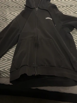 Svart hoodie med dragkedja från Adidas - Säljer en svart hoodie från Adidas med dragkedja framtill och stor vit Adidas-logga på ryggen samt mindre logga på bröstet. Hoodien har huva med dragsko och två stora fickor framtill. Perfekt för en sportig och avslappnad stil.