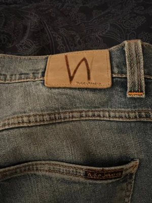 Blå jeans från Nudie Jeans - Säljer ett par klassiska blå jeans från Nudie Jeans i 100% ekologisk bomull. Jeansen har en snygg tvätt, bruna kontrastsömmar och Nudie-logga på bakfickan samt läderpatch i midjan. Perfekta för dig som gillar stilrena och hållbara jeans.