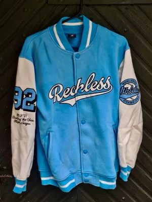 Blå och vit Reckless bomberjacka - Fräsch bomberjacka från Reckless i ljusblått med vita ärmar. Jackan har broderade patches, texten 'Reckless' på bröstet, siffran 92 på ena ärmen och sportiga detaljer. Ribbstickade muddar och krage i blått och vitt. Perfekt för en streetwear-look.