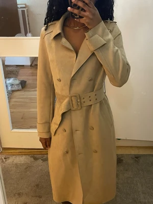Beige trenchcoat med bälte - Stilren beige trenchcoat med klassisk dubbelknäppning och brett bälte i midjan. Jackan har långa ärmar, bred krage och slät yta som ger en elegant look. Perfekt för dig som gillar tidlös och trendig stil.