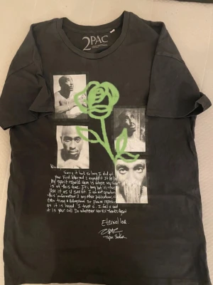 Svart 2PAC t-shirt med tryck - Cool svart t-shirt från 2PAC med fototryck av Tupac Shakur på framsidan, grön ros och vit text. T-shirten har rund hals och korta ärmar. Perfekt för dig som gillar streetwear och vill ha en unik look.