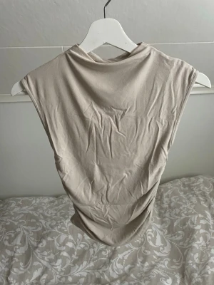 Beige ärmlös topp  - Snygg beige topp i mjukt material med rynkade sidor som ger en cool effekt. Modellen är ärmlös och har en hög halsringning. 