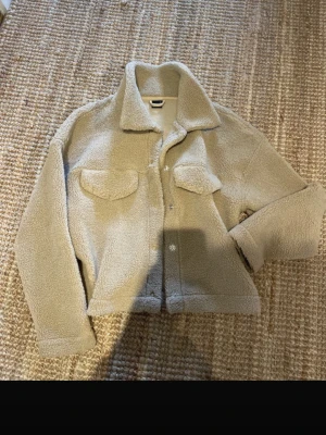 Beige teddyjacka med fickor - Mysig beige teddyjacka i fluffigt material med krage och två bröstfickor med lock. Jackan har knäppning framtill och en avslappnad passform. Perfekt för kalla dagar när du vill vara både varm och trendig.