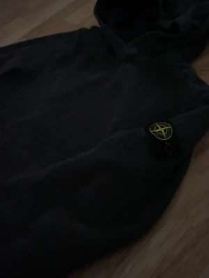 Svart hoodie från Stone Island - Säljer en svart hoodie från Stone Island med den klassiska loggan på vänster ärm. Hoodien har huva och ribbade muddar vid ärmslut och nederkant. Perfekt för dig som gillar streetwear och vill ha en stilren look.