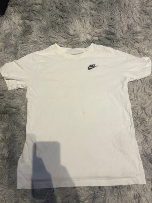 Nike T-shirt - Klassisk vit T-shirt från Nike med svart logga på bröstet. Tillverkad i mjuk bomull och har en normal passform med korta ärmar. Perfekt till vardags för en clean och sportig look. XL i barn storlek