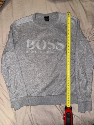 Grå sweatshirt från Hugo Boss - Snygg grå sweatshirt från Hugo Boss med stort BOSS-tryck på bröstet. Tröjan har rund halsringning, långa ärmar och ribbade muddar vid ärmslut och nederkant. Perfekt för en avslappnad och stilren look.