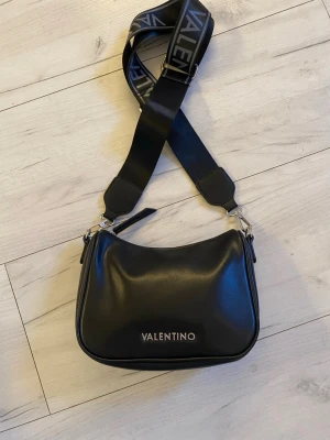 Svart axelväska från Valentino - Snygg svart one sholder bag från Valentino med bred axelrem där loggan står tryckt i vitt. Väskan är i slätt konstläder och har en enkel, stilren design med dragkedja upptill. Som ny. Finns i Bandhagen alt skickad