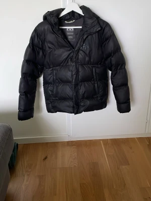 Svart pufferjacka från Helly Hansen - Säljer en svart pufferjacka från Helly Hansen med huva och dragkedja framtill. Jackan har två stora fickor på sidorna och är vadderad för extra värme. Perfekt för kalla vinterdagar och har en klassisk, stilren look. Storlek 152 men passar nog 160 