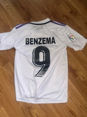 Real Madrid matchtröja Benzema 9 - Säljer en vit Real Madrid matchtröja från Adidas med Benzema och nummer 9 på ryggen. Tröjan har klubbmärke, Adidas-logga och sponsortryck framtill samt La Liga-märke på ärmen. Klassisk krage och lila detaljer på axlarna. Perfekt för fotbollsfans!