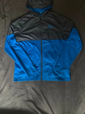Blå och grå vindjacka från Under Armour - Snygg vindjacka från Under Armour i blått och grått med dragkedja framtill. Tillverkad i ett mjukt och sportigt material, perfekt för träning eller chill. Har huva och långa ärmar. Under Armour-logga på bröstet och två sidofickor. Pris kan diskuteras!