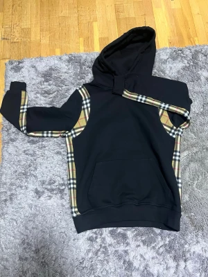 Svart hoodie med rutiga detaljer - Snygg svart hoodie med klassiska beige, rutiga detaljer längs ärmar och sidor. Hoodien har en stor ficka framtill och en rymlig huva. Hoodien saknar snören. Perfekt för dig som vill ha en streetwear-look med lite extra stil.