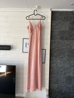 Ljusrosa långklänning i satin - Elegant ljusrosa långklänning med smala axelband och v-ringad urringning. Klänningen har en mjuk, glansig satinfinish och figurnära passform som ger en stilren siluett. Perfekt för sommarens speciella tillfällen. Fläckar syns på bild 3