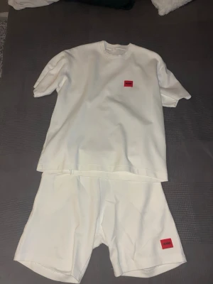 Vitt set från Hugo med röd logga - Säljer ett vitt set bestående av en t-shirt och shorts. Båda plaggen har en röd logga framtill. Setet har en enkel och stilren design med rund halsringning på t-shirten och resår i midjan på shortsen. Perfekt för en avslappnad look.