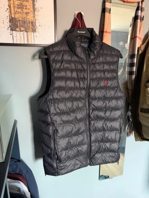 Svart dunväst från Polo Ralph Lauren - Snygg svart dunväst från Polo Ralph Lauren med klassisk quiltad design och dragkedja framtill. Västen har hög krage och den ikoniska röda loggan broderad på bröstet. Perfekt att slänga över en hoodie eller tröja för extra värme.