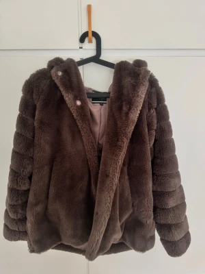 Brun fuskpälsjacka med huva - Mysig brun fuskpälsjacka med huva och breda, horisontella paneler på ärmar och rygg. Jackan har en mjuk insida i ljusare tyg och stängs med tryckknappar framtill. Perfekt för kalla vinterdagar och ger en lyxig känsla till din outfit.