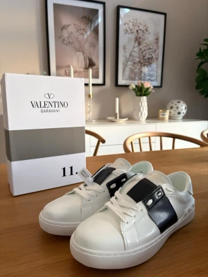 Valentino Garavani vita sneakers - Snygga vita sneakers från Valentino Garavani med bred svart läderdetalj och nitar på sidan. Klassisk låg modell med vita skosnören och rund tå. Tillverkade i slätt läder för en lyxig känsla och stilren look.