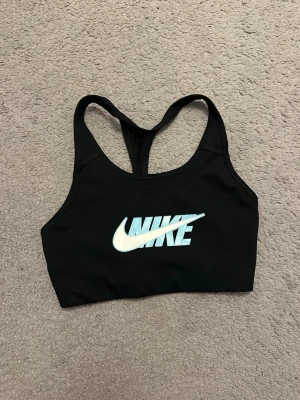 Svart sport-bh från Nike - Snygg svart sport-bh från Nike med klassisk logga i vitt och ljusblått framtill. Bh:n har brottarrygg och är tillverkad i ett stretchigt funktionsmaterial som passar perfekt för träning. Enkel och stilren design med bra stöd.