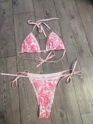 Rosa och vit bikini med knyt - Söt bikini i rosa och vitt med blommigt mönster. Triangeltopp och trosa med knyt i sidorna och nacken för justerbar passform. Hör av dig om du har frågor🥰