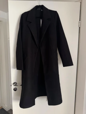 Svart lång kappa med knytning - Stilren svart lång kappa med klassisk krage och raka ärmar. Kappan har en enkel, tidlös design och två sidofickor. Perfekt för dig som vill ha en minimalistisk och trendig look.