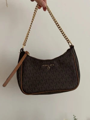 Michael Kors Jet Set Carm Bag - Snygg brun handväska från Michael Kors med guldiga kedjedetaljer och dragkedja. Väskan har MK-logga och mönster över hela ytan samt bruna läderdetaljer längs kanterna. Perfekt storlek för det viktigaste och ger en lyxig känsla.