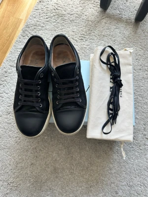 Svarta sneakers från Lanvin - KAN TRÄFFAS I STHLM FÖR SNABB AFFÄR! Stilrena svarta sneakers från Lanvin med ovandel i mocka och läder, samt tåparti i slätt läder. Skorna har svarta skosnören, vita sulor och diskreta metalldetaljer vid snörningen. Medföljer extra skosnören och dustbag.