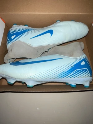 Nike vita och blå fotbollsskor - Säljer ett par ljusblåa  och mörk blå Nike fotbollsskor med stilren design och högt skaft. Skorna har blå detaljer och Nike-logga på sidan, samt elastisk mudd för skön passform. Perfekta för dig som vill ha snygga och funktionella skor på planen.