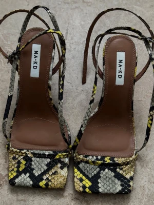 NA-KD sandaler med ormmönster - Snygga sandaler från NA-KD med trendigt ormmönster i beige, svart och gul. Skorna har fyrkantig tå, smala remmar och klack. Materialet är konstläder med struktur. Perfekta för dig som vill sticka ut med coola detaljer och modern design.