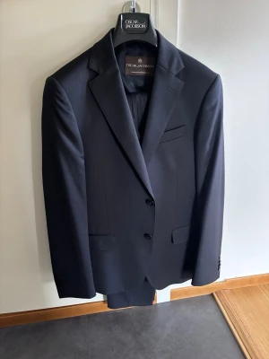 Tredelad kostym från Oscar Jacobson - kavaj, väst och byxor - Oscar Jacobson kostym i tre delar, bestående av en kavaj, en väst och byxor. Kavajen är av modellen Edmund Blazer, västen är Carlo Waistcoat och byxorna är Damien Trousers. Alla delar är i samma färg och stil, vilket skapar en enhetlig och elegant look. Kostymen levereras med en tillhörande plaggpåse från Oscar Jacobson.  Endast använd en gång 