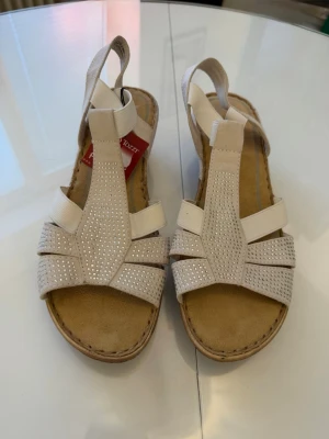 Nya beige sandaler med strassdetaljer - Snygga beige sandaler från Rieker med elastiska band och glittrande strassdetaljer på remmarna. Sköna att bära tack vare mjuk innersula och flexibel sula. Perfekta för dig som vill ha både stil och komfort i sommar.