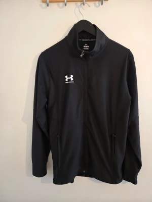 Svart träningsjacka från Under Armour - Svart träningsjacka från Under Armour med hel dragkedja och ståkrage. Jackan har två fickor med dragkedja och en vit logotyp på bröstet. Perfekt för träning eller uppvärmning. Tillverkad i ett lätt och stretchigt material.