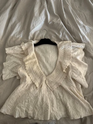 Otroligt fin blus volang Zara - Romantisk vit blus i spets med bred volangkrage och korta ärmar. Blusen har en djup v-ringning och vackra spetsdetaljer som ger en vintagekänsla. Perfekt för dig som gillar en feminin och trendig stil.