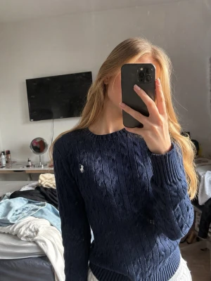 Navy cableknit från Ralph Lauren - Kabelstickad mörkblå tröja från Ralph Lauren Sport i 100% bomull. Klassisk rund halsringning och vit broderad logga på bröstet. Tröjan har ribbade muddar vid ärmslut och nederkant, perfekt för en stilren och tidlös look.