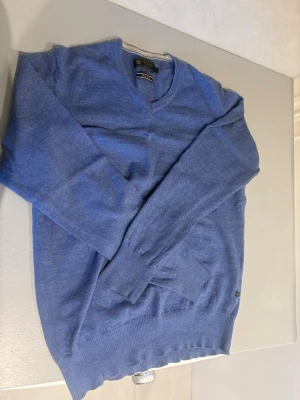 Blå merinoulltröja från Riley - Snygg blå stickad tröja från Riley i mjuk och tunn merinoull. Tröjan har V-ringning, långa ärmar och ribbade muddar vid ärmslut och nederkant. Perfekt för lager-på-lager och passar till många olika stilar.