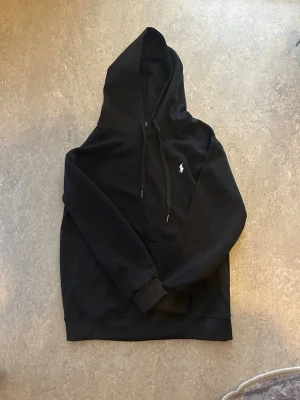 Svart hoodie från Polo Ralph Lauren - Säljer en svart hoodie från Polo Ralph Lauren med klassisk huva och dragsnören. Tröjan har en stor ficka framtill och den ikoniska lilla vita Polo-loggan på bröstet. Tillverkad i mjukt bomullsmaterial som är skönt att ha på sig.