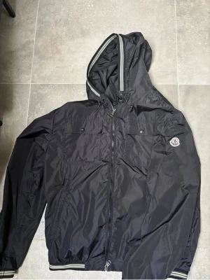 Moncler Windbreaker 4 - Svart lättviktsjacka från Moncler med huva och klassisk logga på ärmen. Stilren och praktisk modell med dragkedja framtill, två bröstfickor med knapp. Perfekt som vår-/sommarjacka.  Jackan är i fint skick och passar både till vardag och en mer uppklädd stil. Storlek 4 vilket motsvarar M/L