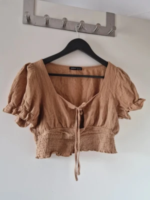 Beige croppad blus med puffärm - Söt beige croppad blus från Shein med puffärmar och smockad midja. Toppen har v-ringning och knyt framtill, vilket ger en avslappnad men trendig look. Välanvänd men den är fortfarande i fint begagnat skick. Katt finns i hemmet