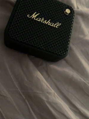 Marshall bärbar högtalare svart - Snygg och kompakt bärbar högtalare från Marshall i svart med guldiga detaljer. Högtalaren har en robust design med texturerad yta och klassisk Marshall-logga framtill. Perfekt för att ta med sig musiken överallt. Låter jättebra Inga problem alls. Laddare ingår inte . Men det funkar med en vanlig dator laddare. Låter jätte bra.