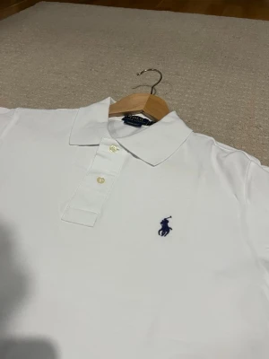 Vit pikétröja, Ralph Lauren - Klassisk vit pikétröja från Ralph Lauren med kort ärm och broderad marinblå logga på bröstet. Tröjan har krage, knappar framtill och är tillverkad i bomull. Perfekt för en stilren och tidlös look.