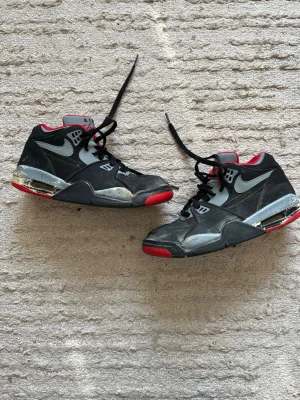 Nike Air Flight svart/grå sneakers - Säljer ett par klassiska Nike Air sneakers i svart och grått med röda detaljer. Skorna har hög modell, synlig Air-sula och ikonisk Swoosh på sidan. Materialet är en mix av läder och syntet. Perfekta för dig som gillar retrostil och streetwear.
