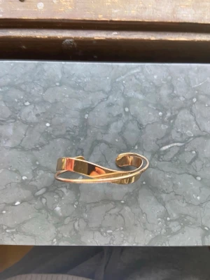 Guldigt öppet armband i metall - Snyggt armband i guldfärgad metall med en öppen design och tvinnad form. Armbandet har en stilren och modern look som passar perfekt till din outfit. Ingen märkning syns på armbandet.
