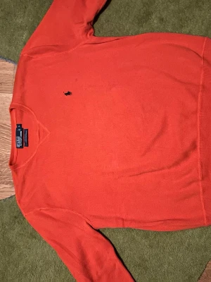 Orange stickad tröja från Polo Ralph Lauren - Snygg orange stickad tröja från Polo Ralph Lauren med klassisk svart logga broderad på bröstet. Tröjan har rund halsringning och långa ärmar. Perfekt för dig som vill ha en färgklick i garderoben.