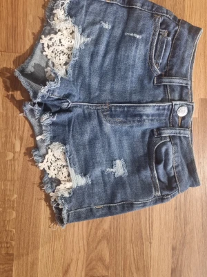 Blå jeansshorts med spetsdetaljer - Snygga blå jeansshorts med slitna detaljer och råa kanter. Shortsen har vita spetsdetaljer. Storlek W24 