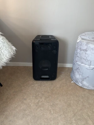 Stor svart bärbar Bluetooth-högtalare - Snygg svart bärbar högtalare med kraftfullt ljud och inbyggd belysning som lyser blått. Högtalaren har en robust rektangulär form och är tillverkad i hård plast med metallgaller på framsidan. Perfekt för att spela musik trådlöst via Bluetooth.