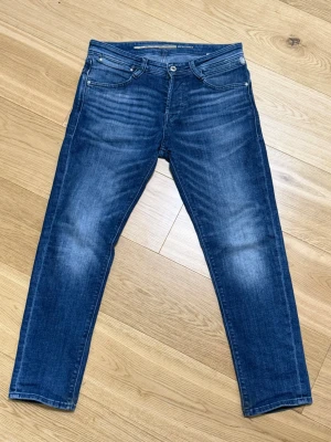 Blå jeans från J&J - Snygga blå jeans från J&J Jeans med klassisk femficksdesign och gul söm på bakfickorna. Slim fit 30/32.