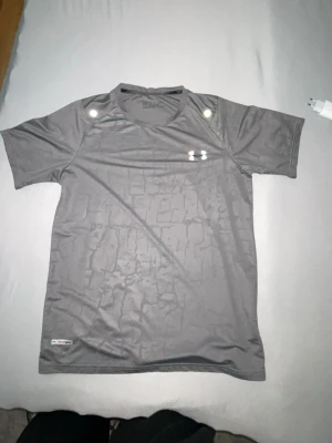 Grå träningströja från Under Armour - Snygg grå sporttröja från Under Armour med korta ärmar och rund hals. Tillverkad i ett lätt och ventilerande funktionsmaterial, perfekt för träning. Loggan på bröstet har en cool regnbågsfärgad effekt som sticker ut.