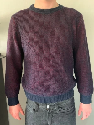 Mörkblå och vinröd stickad tröja - En stilren stickad tröja i mörkblått och vinrött med rund hals och ribbade muddar. Tröjan har en klassisk passform och är perfekt för att hålla värmen. Materialet är mjukt och ger en skön känsla. Passar dig som gillar en enkel men snygg look. Stolek L men mindre i storlek. 